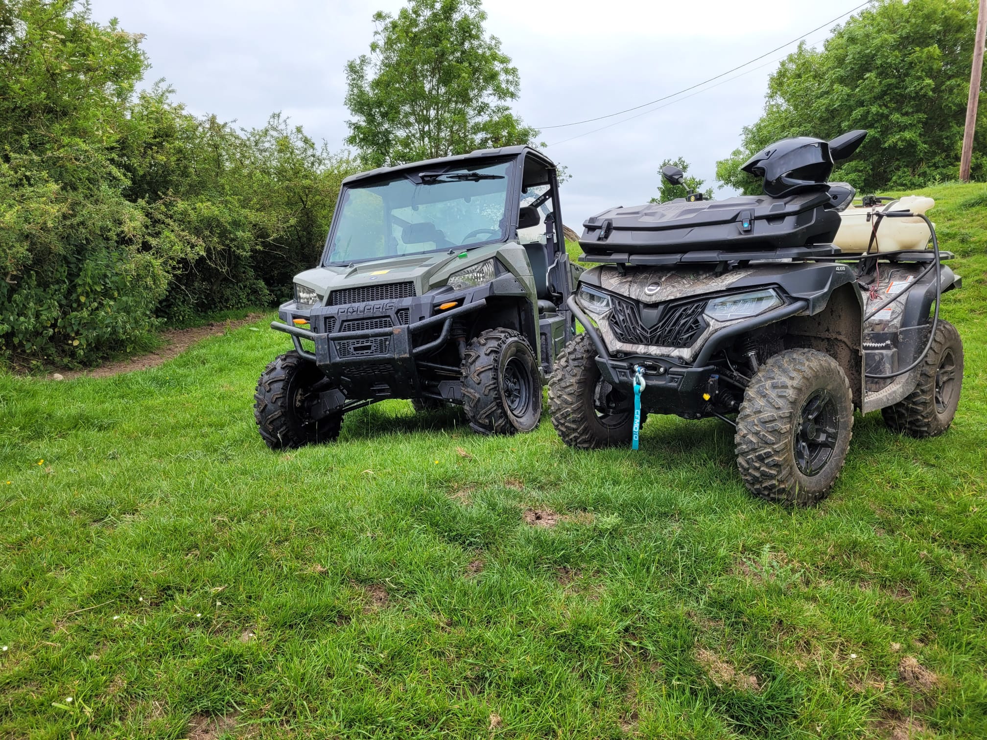 A Polaris utility terrain vehicle and a quad bike with front storage box parked side by side on grass beside trees.