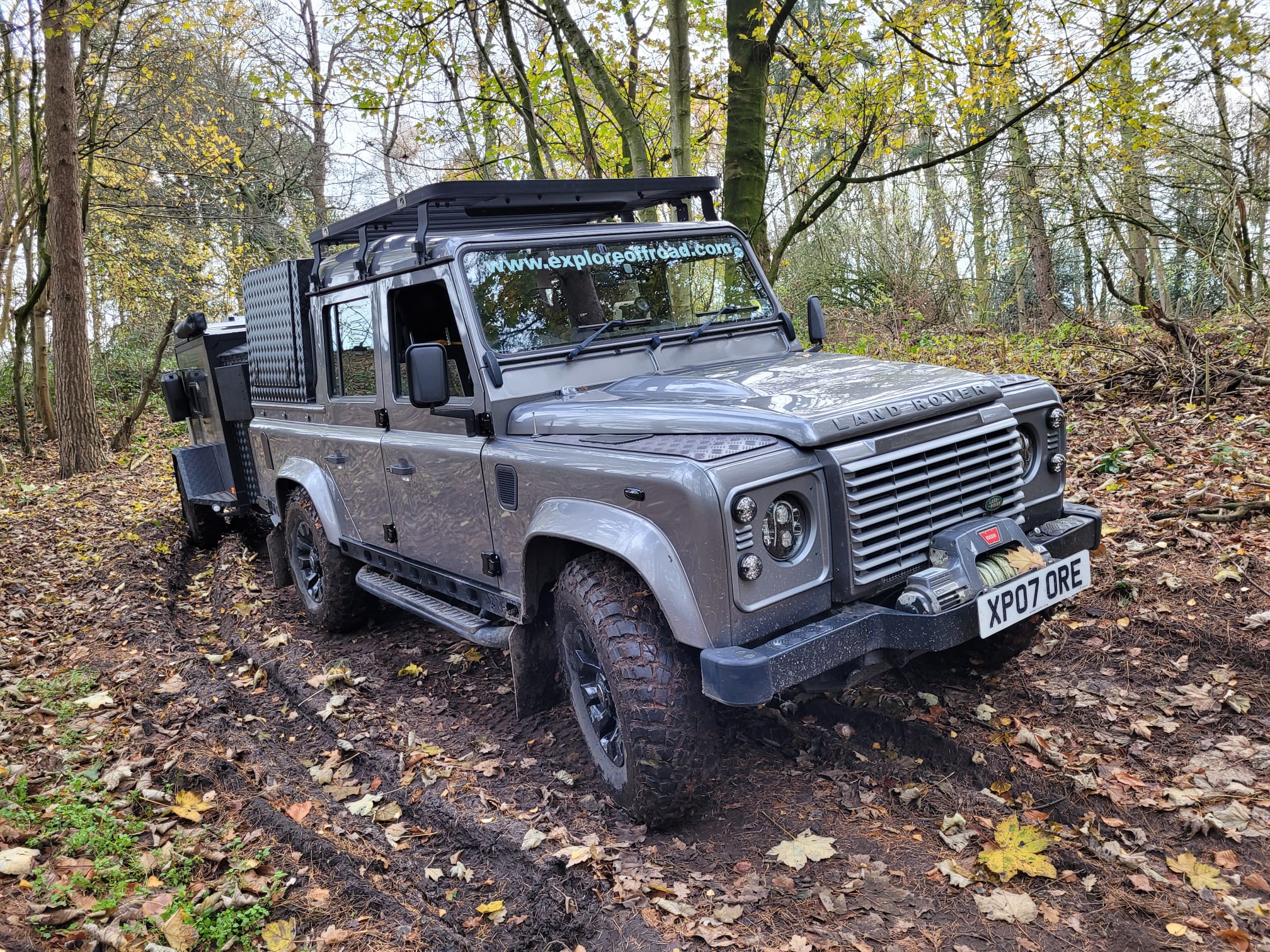 Explore Off-Road grey Land Rover Defender XP07 ORE with roof rack and front winch on a muddy autumn woodland track.
