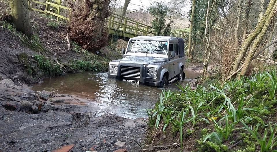 Defender driving through a stream along a woodland route