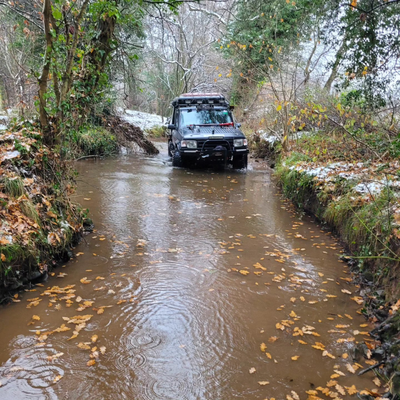 A Land Rover Defender driving through a water-filled woodland track surrounded by trees.